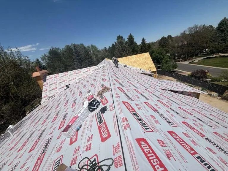 A rooftop covered with synthetic underlayment, tools, and building materials. A person works near the roofs peak. Trees and a street border the construction site. The sky is clear and sunny.