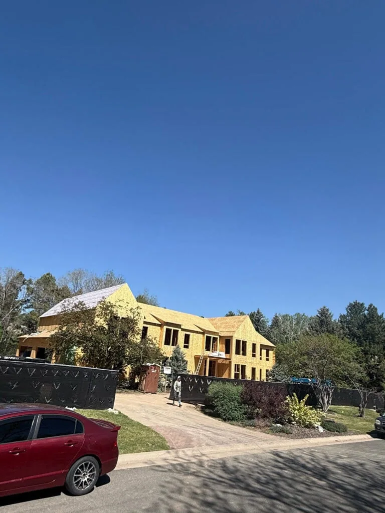 A large house under construction with exposed yellow framing sits behind a black fence. Trees and shrubs surround the property, and cars are parked along the street under a clear blue sky.