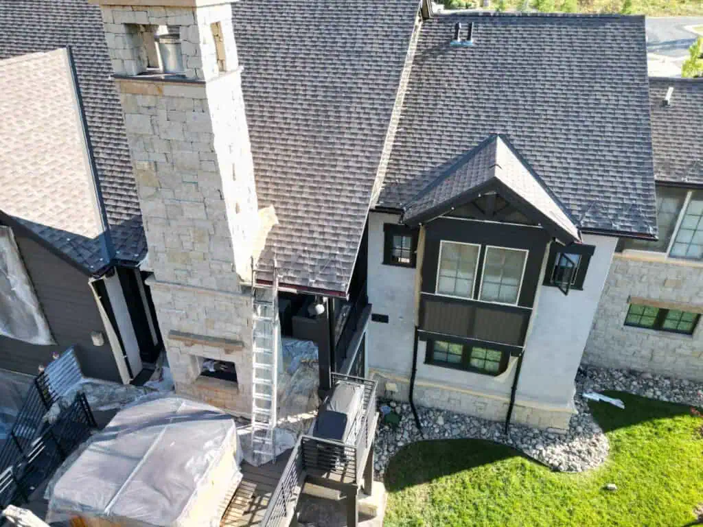 A large modern house with a stone chimney, dark roof shingles, and a metal ladder propped against the roof. The yard has green grass and landscaping rocks along the houses edge.