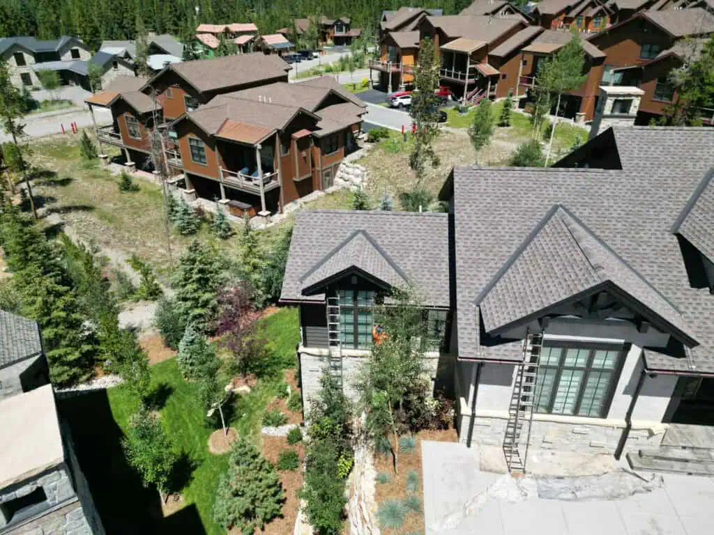 Aerial view of modern mountain homes with brown roofs, surrounded by trees, greenery, and a small road. The houses are close together in a scenic residential neighborhood on a sunny day.