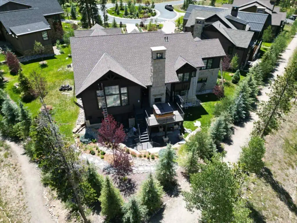 Aerial view of a large modern house with stone and dark wood exterior, surrounded by landscaped greenery, young trees, walking paths, and neighboring homes. A backyard deck is visible at the rear of the house.