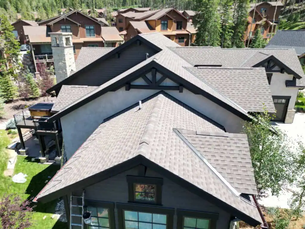 Aerial view of a large, modern house with multiple peaked roofs, surrounded by green trees and neighboring homes in a residential mountain area.