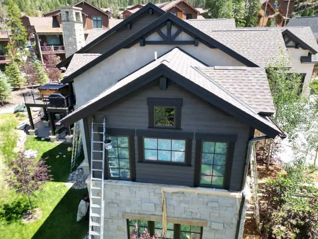 Aerial view of a modern two-story house with stone and dark wood siding, surrounded by green lawn and trees. A ladder leans against the house, indicating ongoing maintenance or painting.