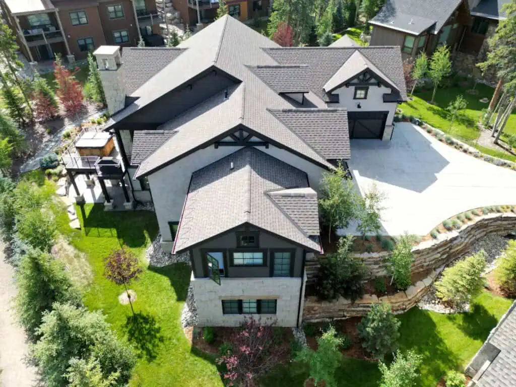 Aerial view of a large, modern house with a dark roof, light exterior walls, and a spacious driveway surrounded by well-maintained landscaping and green trees.