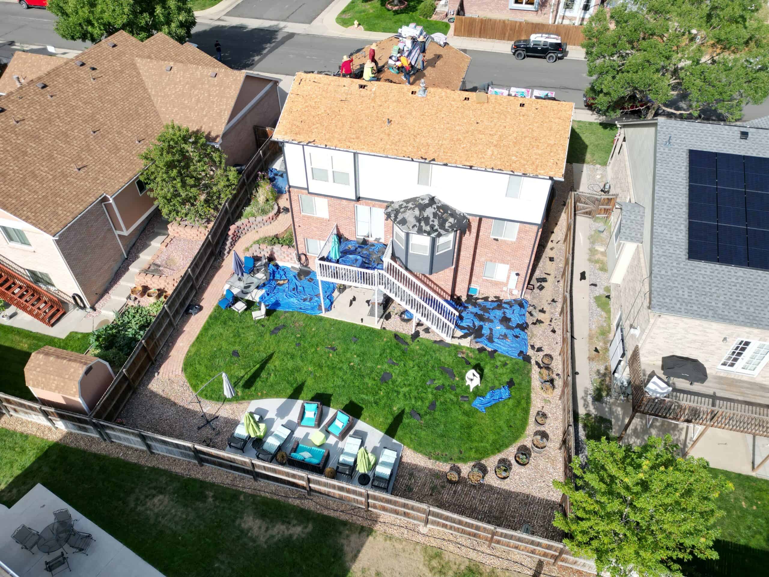 Aerial view of a backyard with green grass and patio furniture. The house has workers on the roof, some covered with tarps, and materials scattered on the ground. Surrounding homes and a street are also visible.