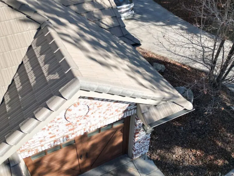 Aerial view of a house roof with tan shingles, a brick wall, brown double garage doors, a leafless tree, and a parked car partially visible on a concrete driveway.