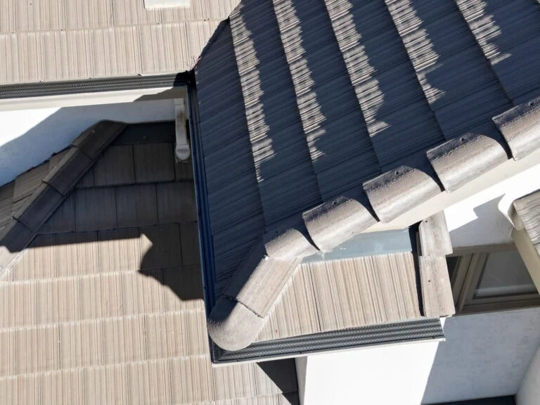 Aerial view of a house roof with overlapping grey tiles, a skylight, and shadows cast by the roof lines. The roof structure creates angular shapes and some window edges are visible on the white exterior wall.