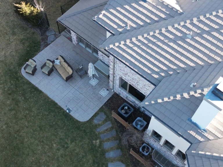 Aerial view of a house’s backyard showing a patio with outdoor furniture, umbrella, and stone pathway, next to the house and grassy area. Rooftop AC units are visible.