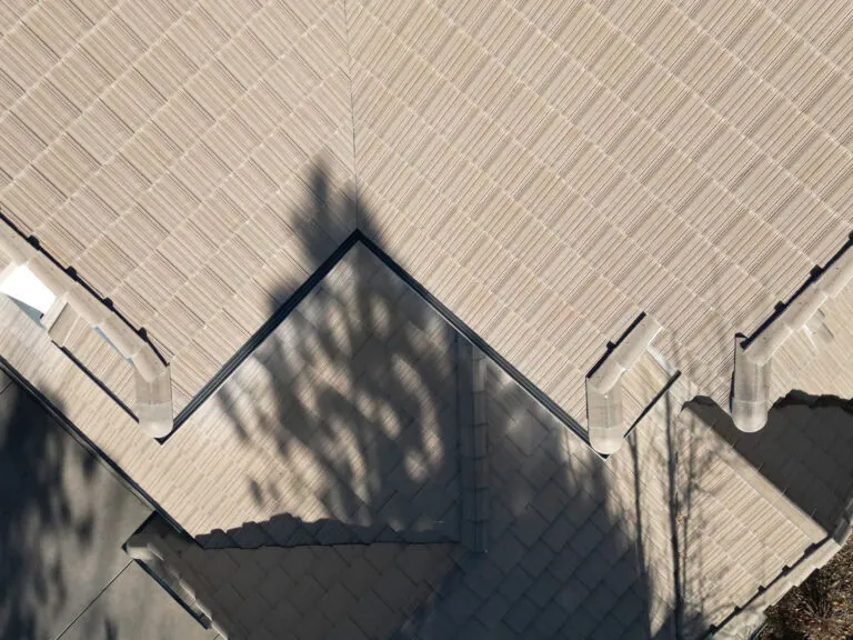 Aerial view of a beige-tiled roof with sharp angles, casting a distinct tree shadow across the surface. Part of a driveway and some foliage are visible along the edges.