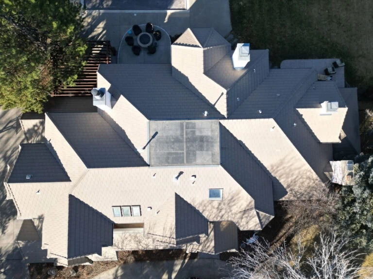 Aerial view of a large light-colored house with multiple roof sections, skylights, chimneys, surrounding trees, patio furniture, and paved driveways. Shadows are cast by trees and parts of the house.