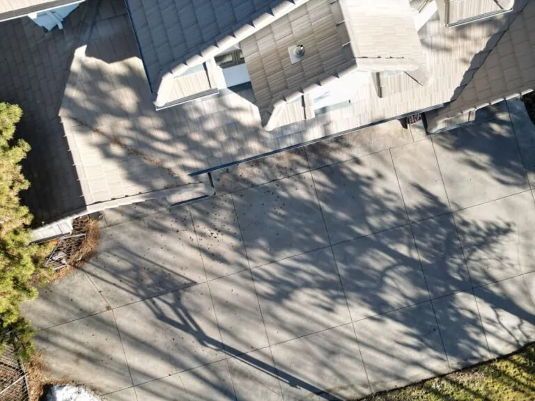Aerial view of a house roof and large concrete driveway, with tree shadows cast across the surface. Some scattered leaves and a bit of snow are visible near the driveways edge.