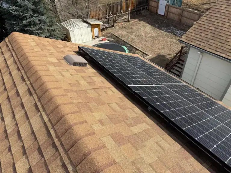 A row of solar panels is installed on the brown shingle roof of a house. In the background, there is a backyard with a shed, trampoline, and dry grass.