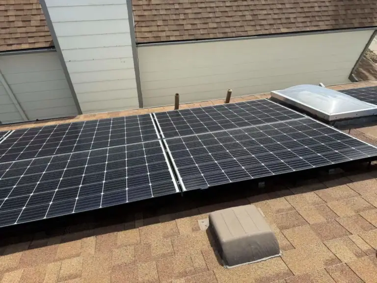 Solar panels installed on a shingled rooftop near a skylight and several roof vents. The background shows sections of light-colored siding from an adjacent building.