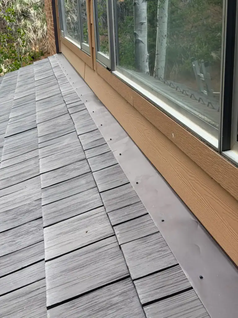 A close-up view of a rooftop with gray shingles and a strip of metal flashing next to large windows reflecting trees and sky.