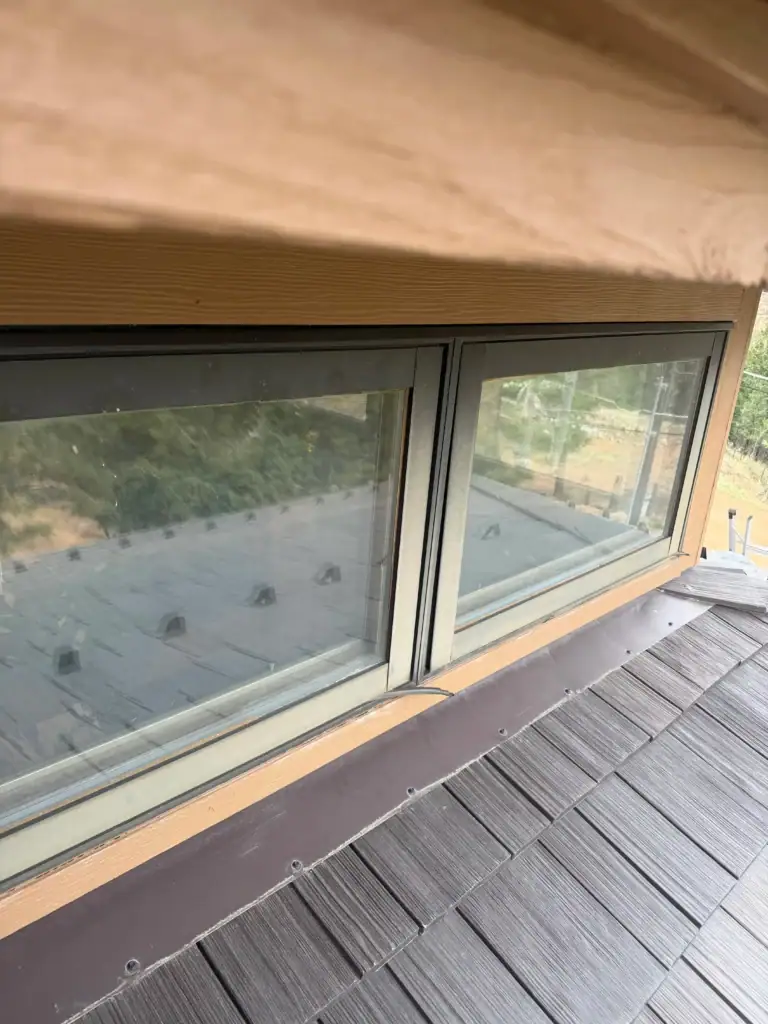 A close-up view of a pair of rectangular windows on a wooden building, reflecting an outside scene. The roof with dark shingles and the edge of the eaves are visible in the foreground.
