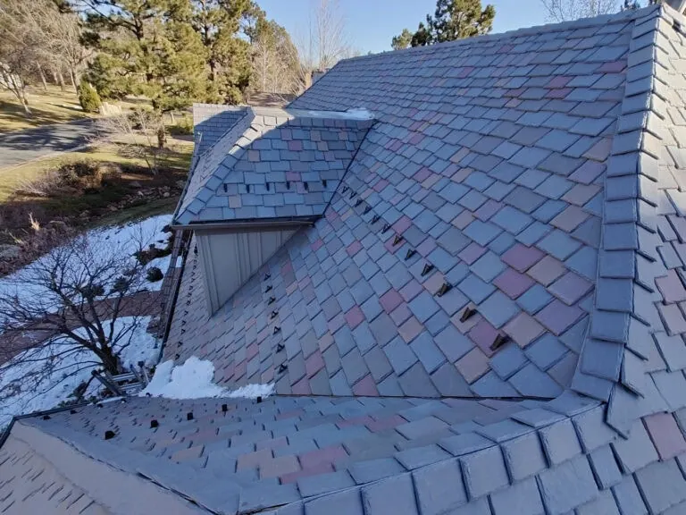 A roof with multicolored shingles, including blue and reddish tones, shows some missing or damaged shingles. There is a small amount of snow in patches on the roof and some snow on the ground below. Trees are in the background.