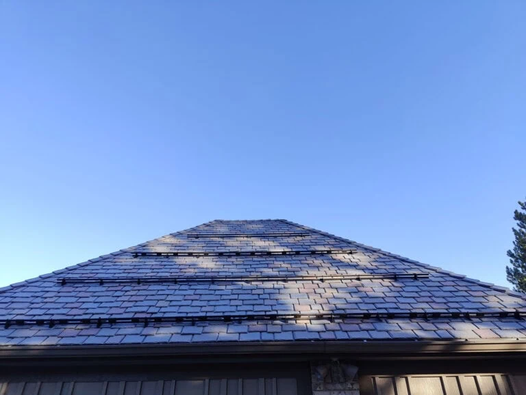 A pyramid-shaped roof with brown shingles is pictured from below under a clear blue sky. Shadows from nearby trees are visible on the roof.
