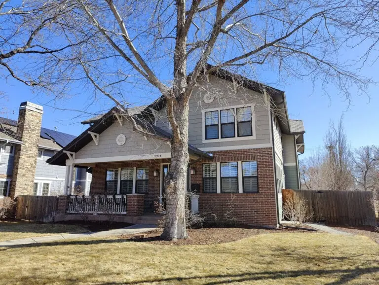 A two-story brick and siding house with black shutters, a covered porch, and a large leafless tree in front, set on a dry, yellowish lawn under a clear blue sky.