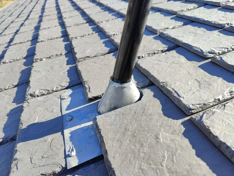 A black pipe protrudes through a gray slate roof, sealed with metal flashing around its base for waterproofing. The sunlight casts shadows across the uneven slate tiles.