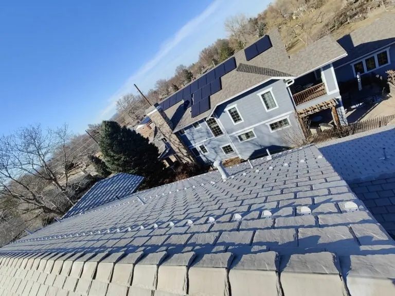 View from a sloped rooftop with shingles, overlooking a blue house with solar panels on the roof, surrounded by trees and a clear blue sky.