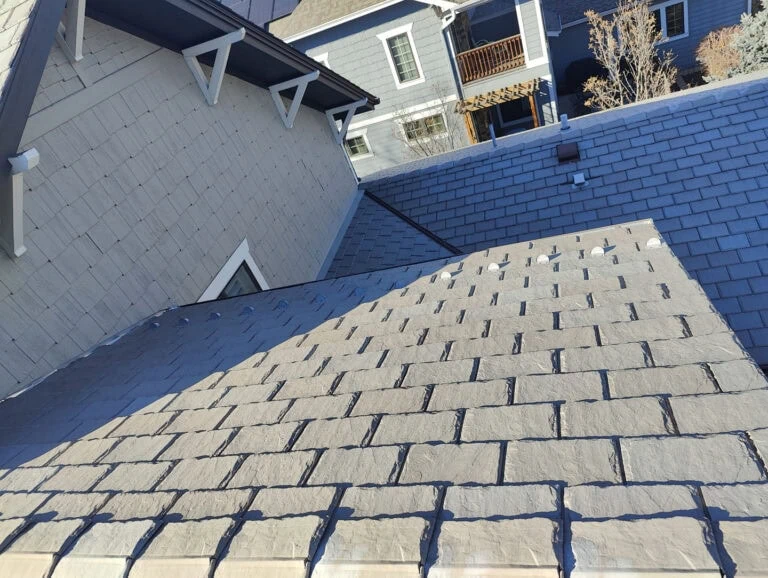 Sunlight casts angular shadows across gray roof shingles of a house. Neighboring houses with blue and gray siding are visible in the background, along with some trees and a clear sky.
