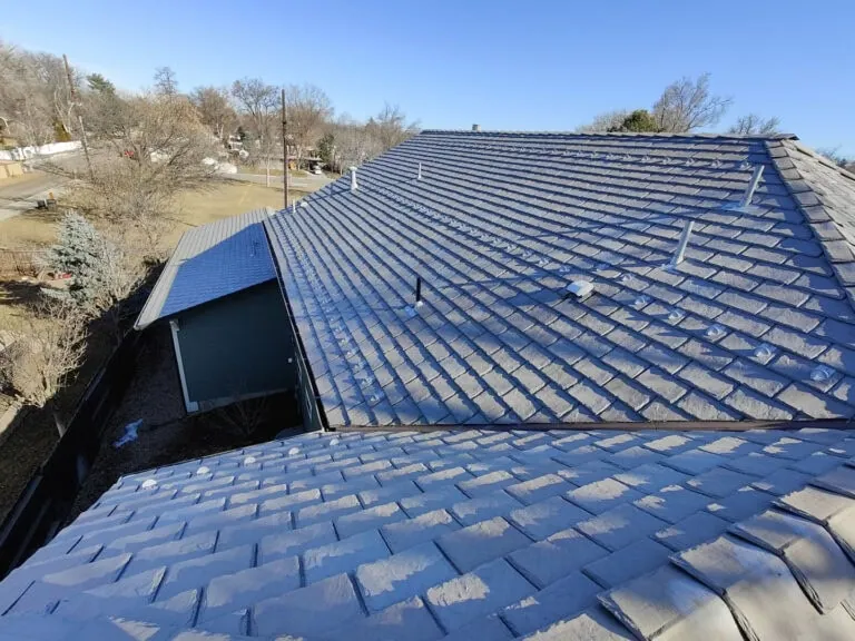 A roof covered in gray shingles is shown under clear blue sky. Vents and pipes protrude from the roof, and trees and houses are visible in the background. Sunlight casts shadows across the shingles.