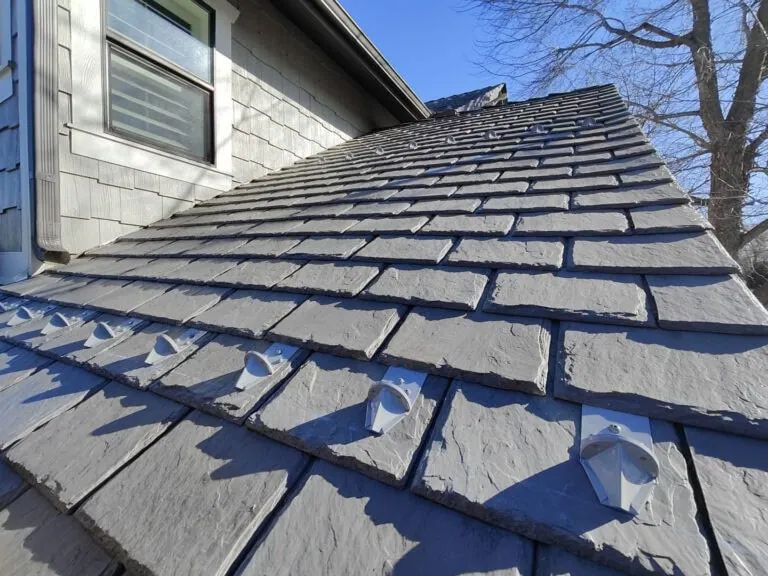 Close-up view of a house’s slate roof with several evenly spaced metal snow guards. The sky is clear and blue, and leafless tree branches are visible in the background.