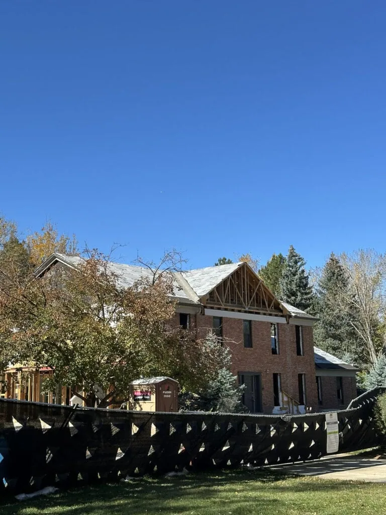 A two-story brick house under construction, with exposed roof trusses and windows missing. Construction fencing surrounds the site, and trees with autumn foliage are visible under a clear blue sky.