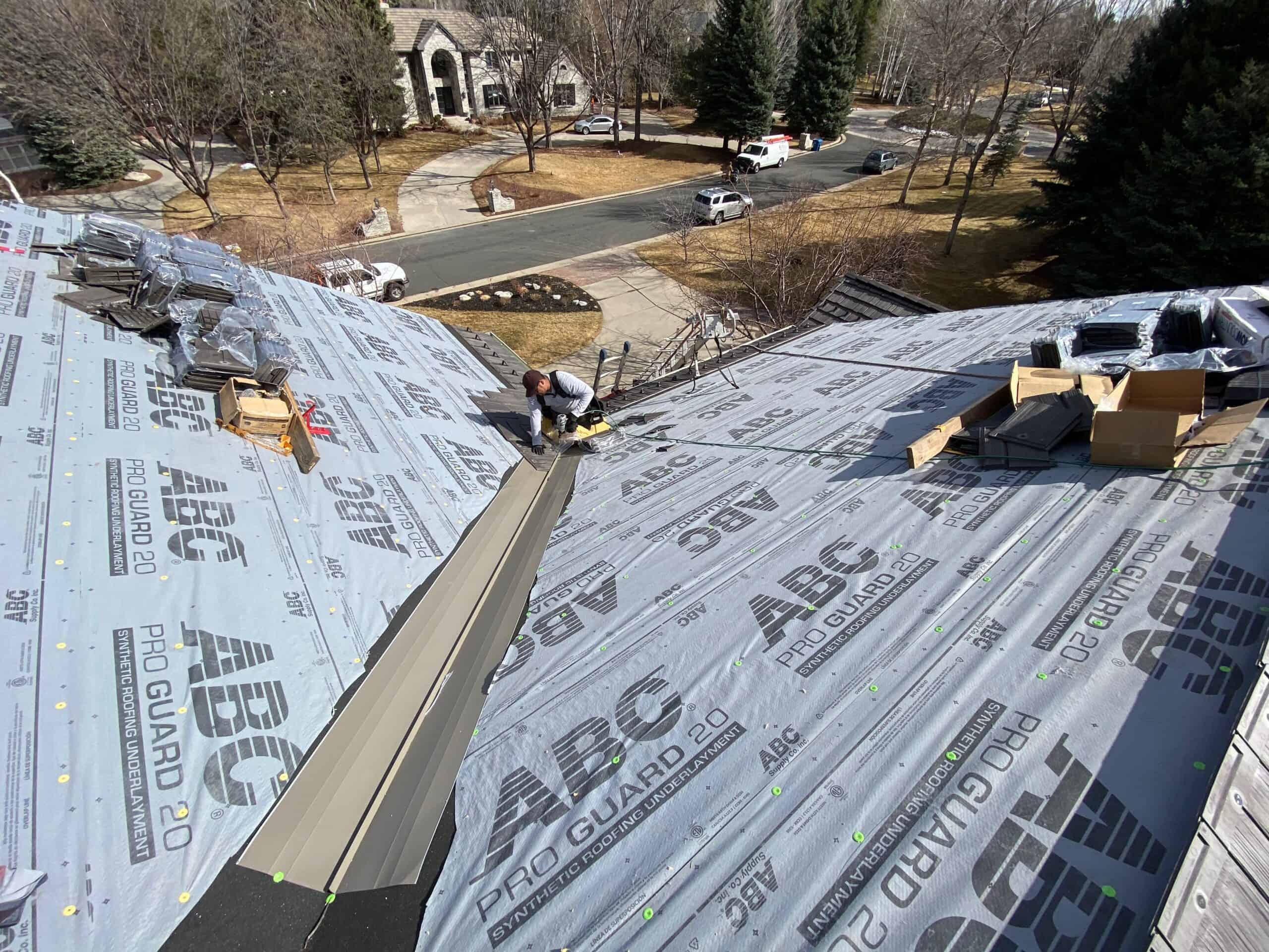 A worker kneels on a house roof covered with waterproof underlayment, installing metal roofing panels. Tools, materials, and boxes are scattered around. Trees, cars, and houses are visible in the background on a sunny day.