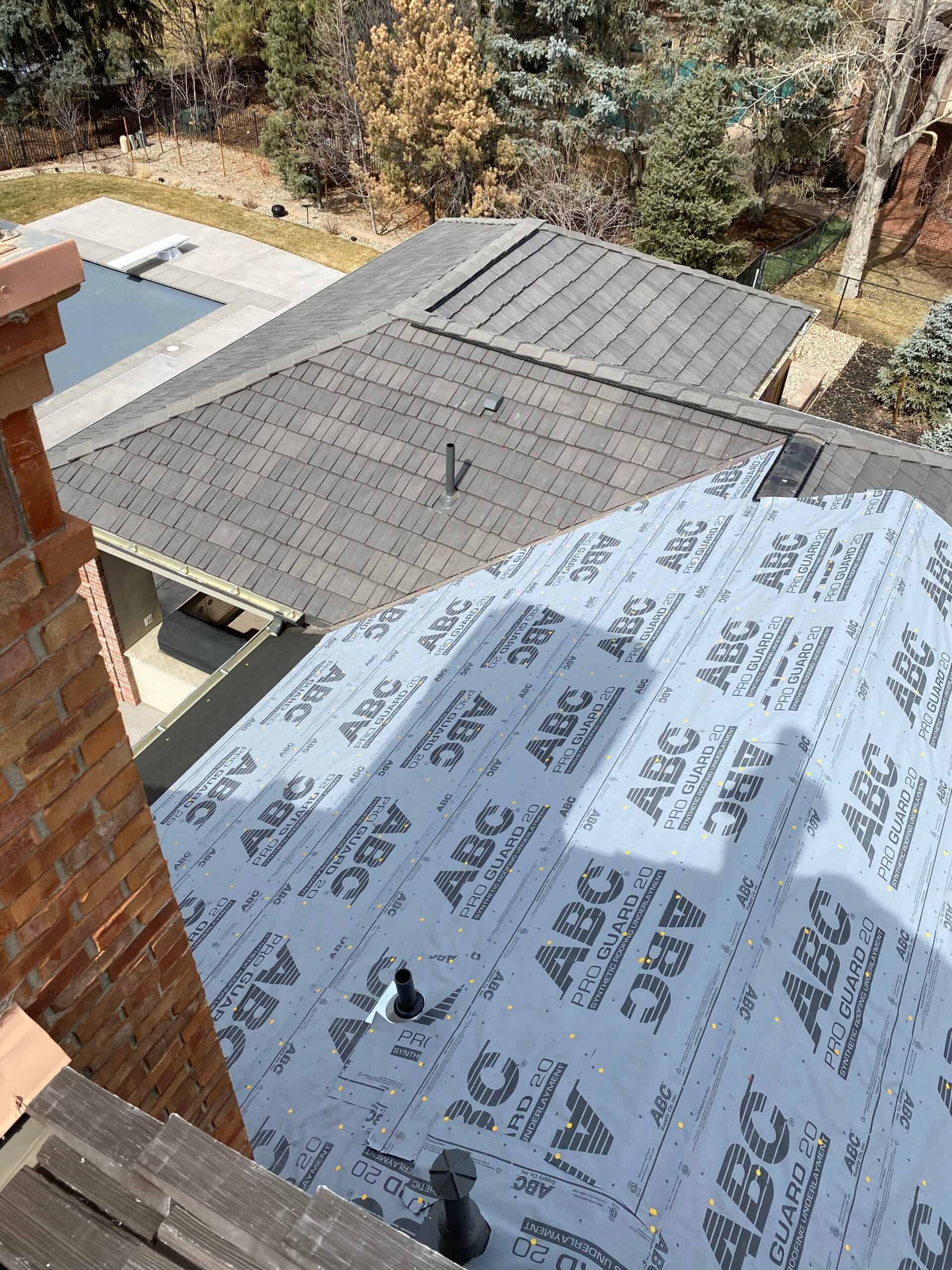View from a rooftop showing part of a roof covered in synthetic underlayment, labeled “ABC,” next to an area with installed shingles. Trees, a pool, and a patio are visible in the background.