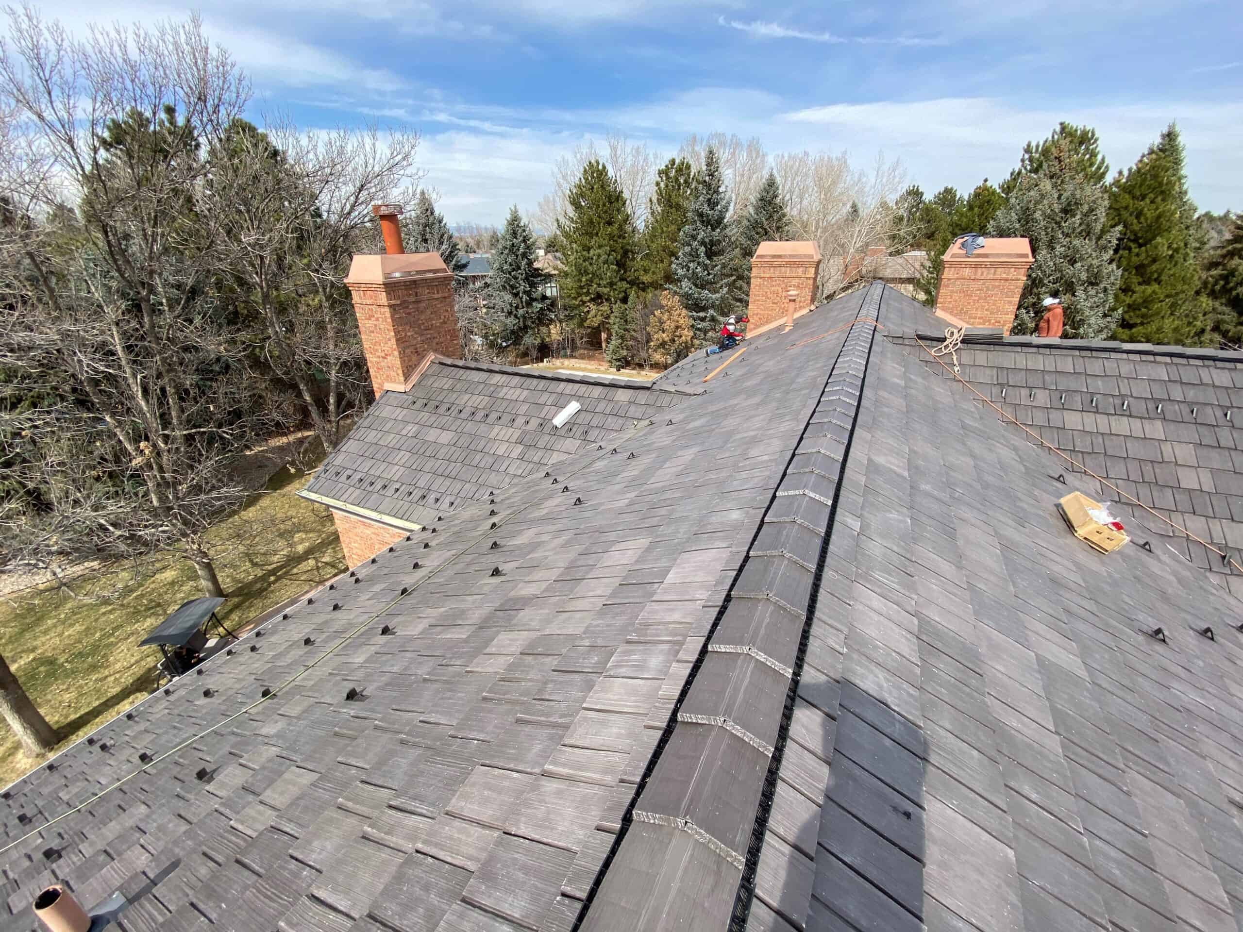 View of a sloped gray shingle roof with multiple brick chimneys, surrounded by tall trees and a grassy yard under a partly cloudy sky. Roofing materials and tools are visible on the roof.