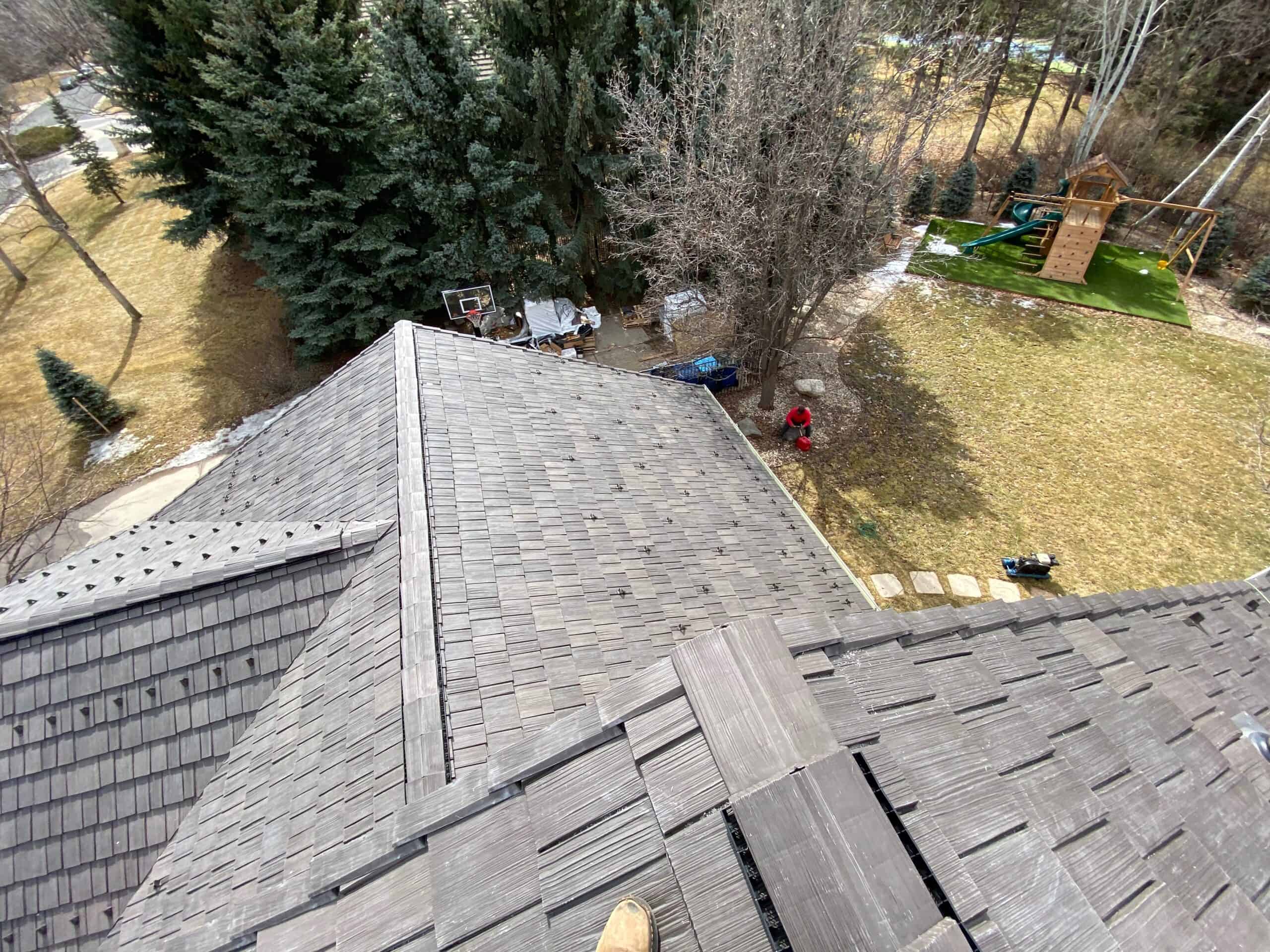 View from a rooftop looking down at a backyard with scattered trees, a playground set, some equipment, and a person standing on the grass. The photo is taken from a high, steep angle.