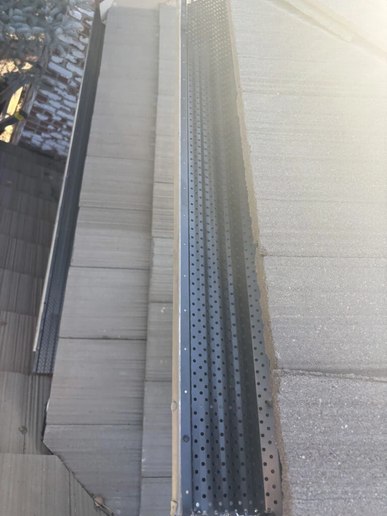 A close-up view of a building’s rooftop edge featuring grey roof tiles and a black metal gutter guard with small, evenly spaced holes. Sunlight is partially visible on the shingles.