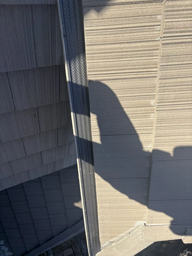 A shadow of a person is cast over a tiled roof with overlapping shingles and a black, perforated metal strip running along the ridge.