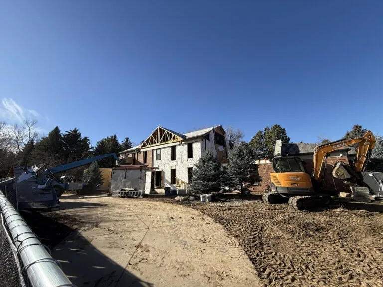 A house under construction with exposed framing, surrounded by construction equipment and dirt, under a clear blue sky. Pine trees and other greenery are visible in the background.
