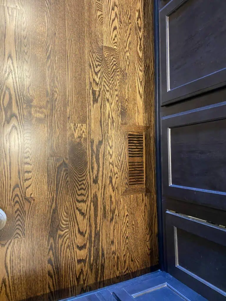 Wooden floor with a rectangular floor vent near a dark wooden door. The floor has a natural wood grain pattern and the door has decorative panels.