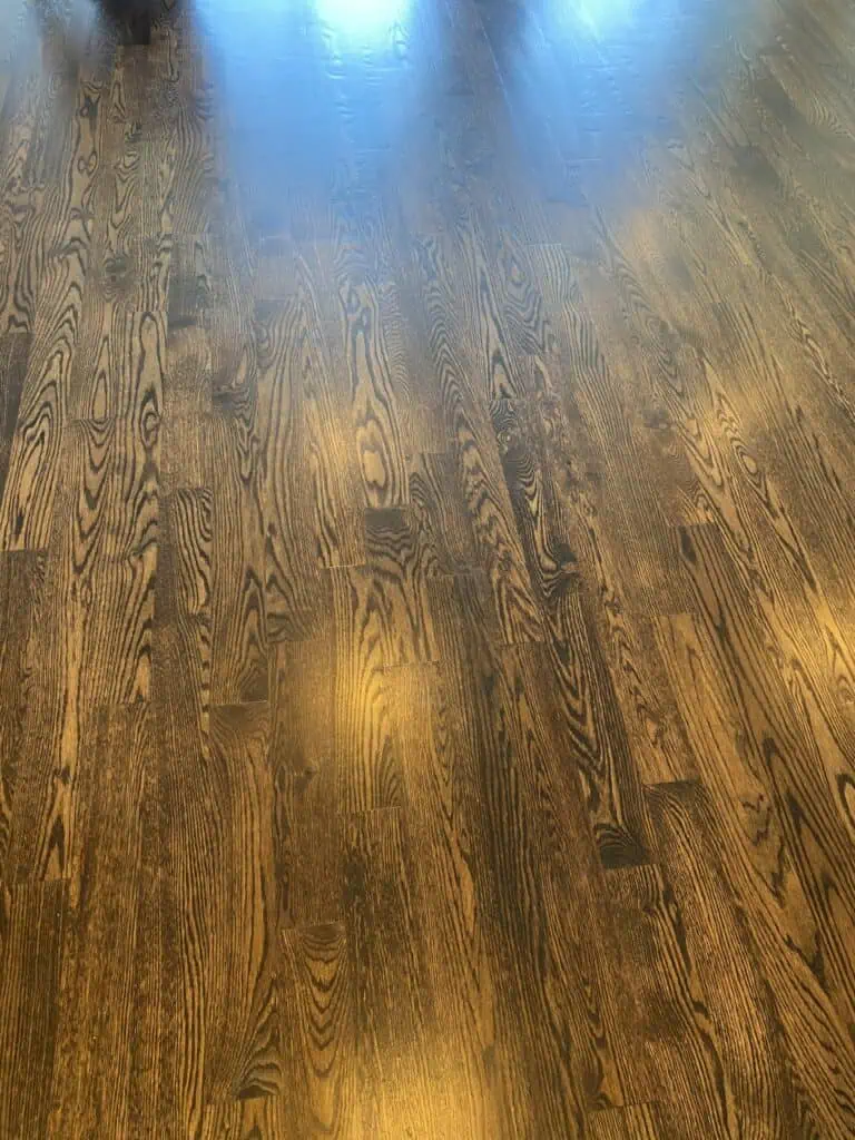 A close-up of a polished, dark wood floor with visible natural grain patterns and some light reflections across the surface.