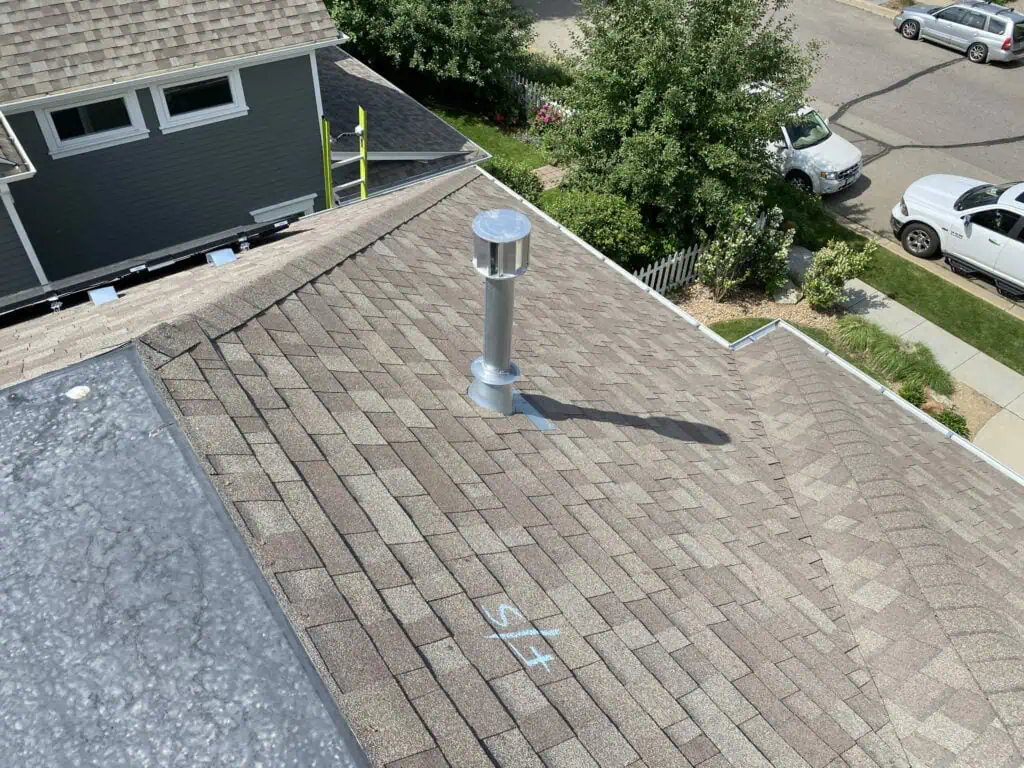 A rooftop with brown shingles features a metal vent pipe. There is a chalk marking with L/S on the roof. Nearby are a ladder, a house, trees, and parked cars on the street.