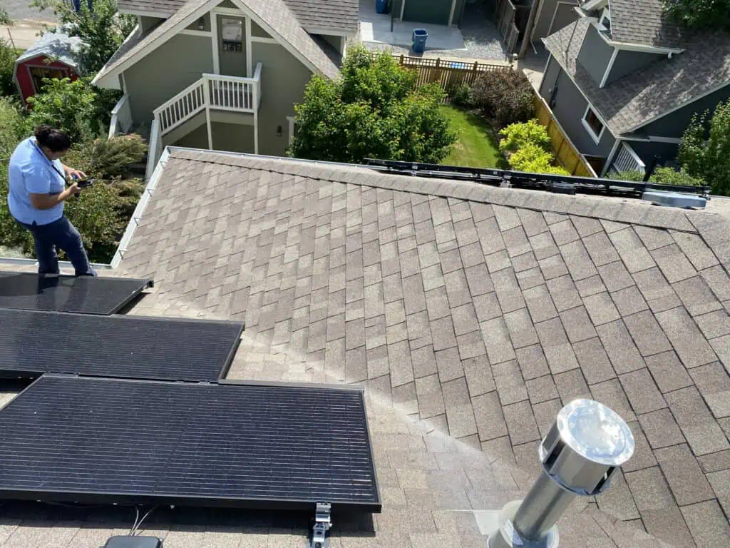 A person stands on a shingled roof near solar panels, holding a device. Several houses with green trees and a driveway are visible in the background.