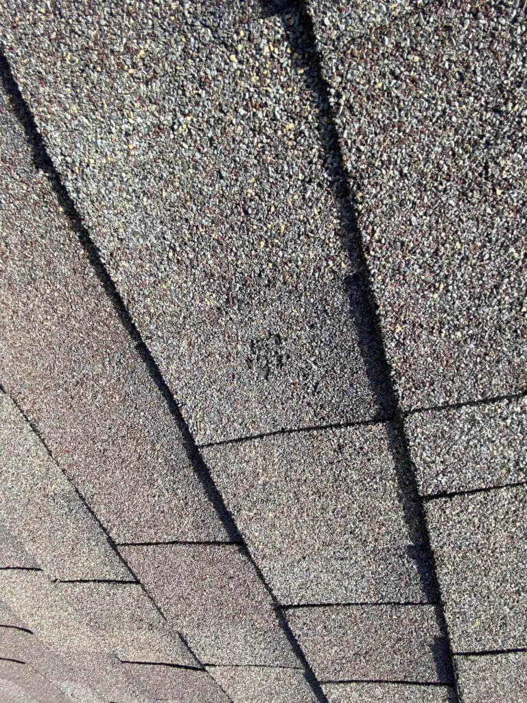 Close-up of brown asphalt roof shingles, showing the texture and overlapping pattern. There is a small black spot or stain near the center of the image.