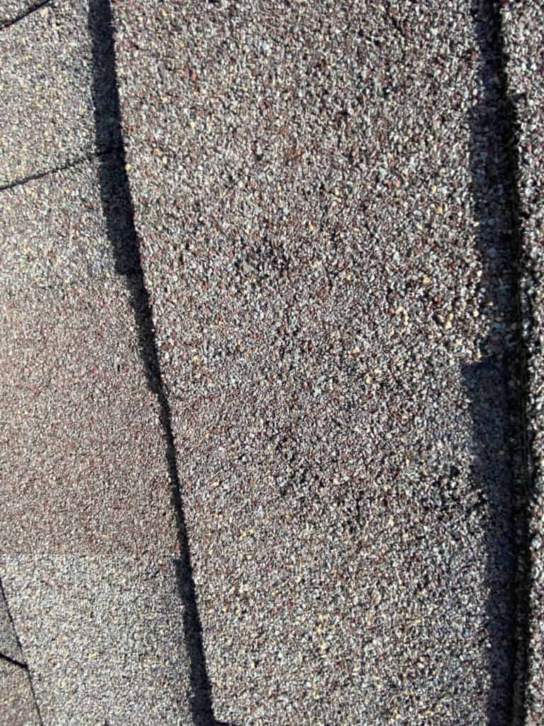 Close-up photo of overlapping brown asphalt roof shingles, showing their rough, granulated texture and shadows created by sunlight.