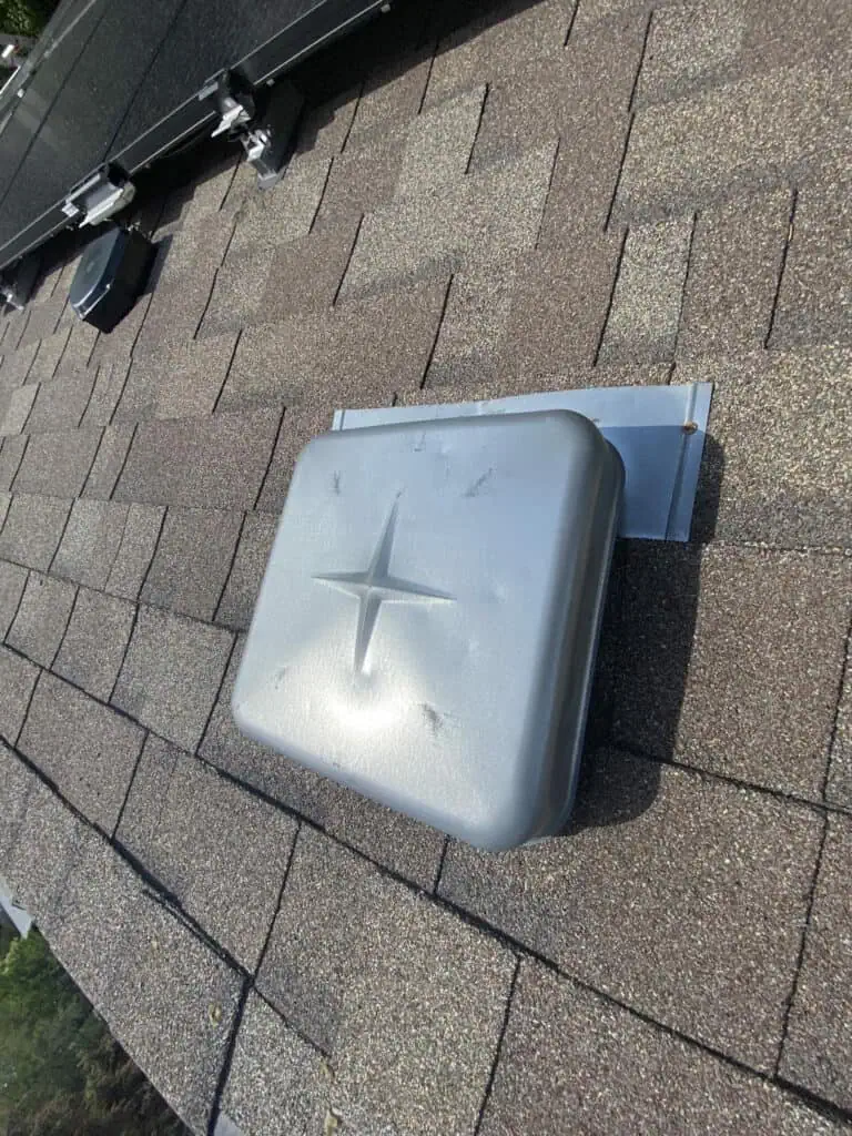 A metal vent cap with a cross-shaped indentation is mounted on a shingled roof. Solar panel mounts are partially visible in the top left corner.