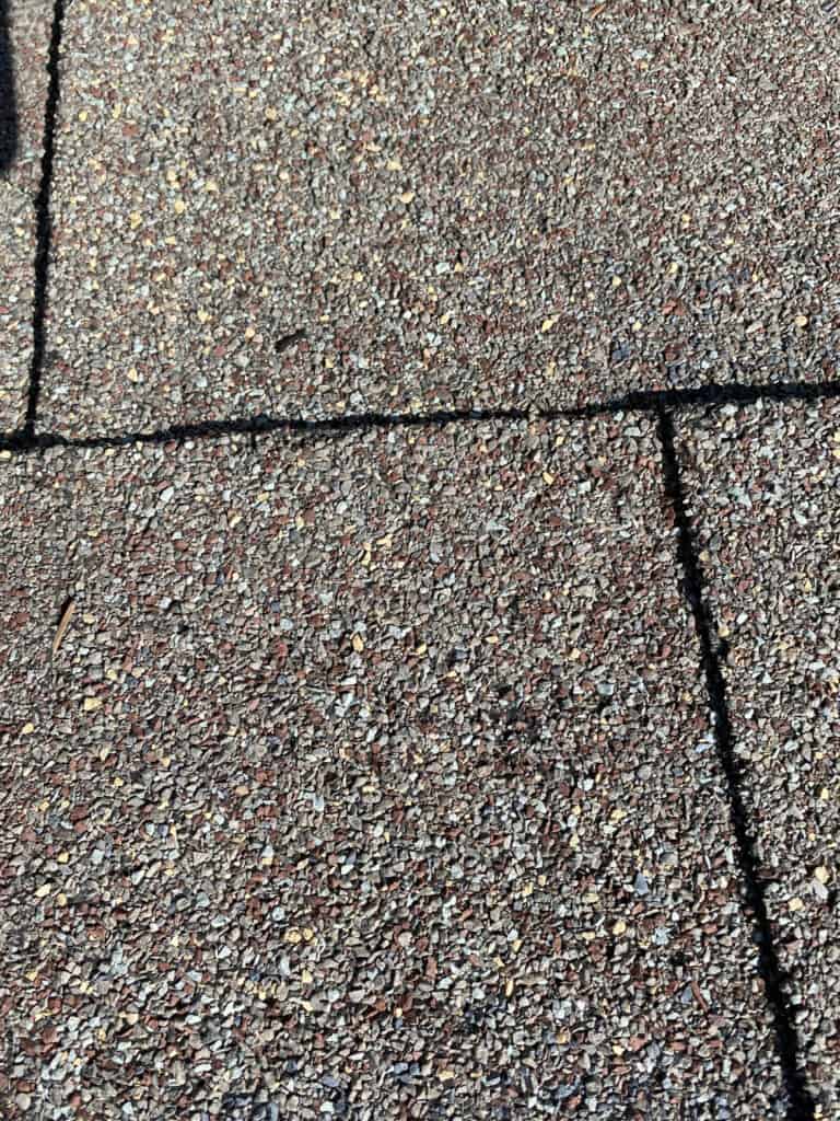 Close-up of asphalt roof shingles with a visible horizontal and vertical seam, showing their rough, gritty texture in shades of brown, gray, and tan under sunlight.