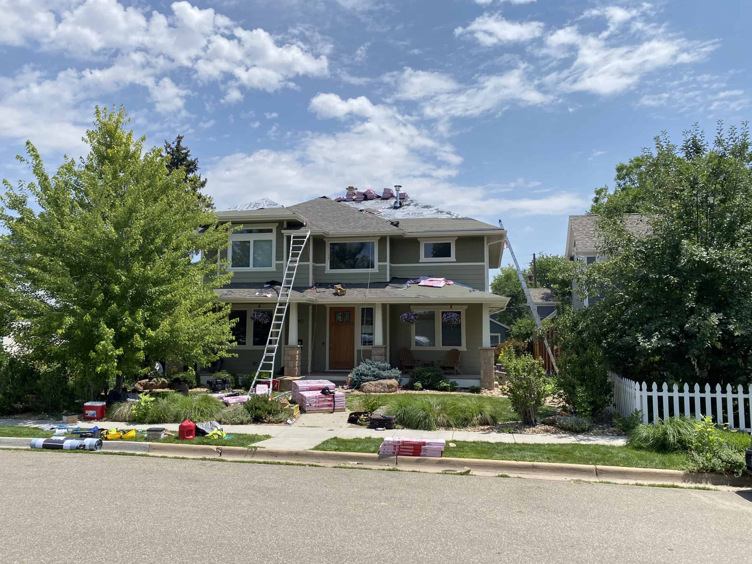 A two-story house with roofing materials on the roof, a ladder leaning against it, and supplies scattered on the lawn, indicating roof work is in progress. The sky is partly cloudy and trees surround the house.