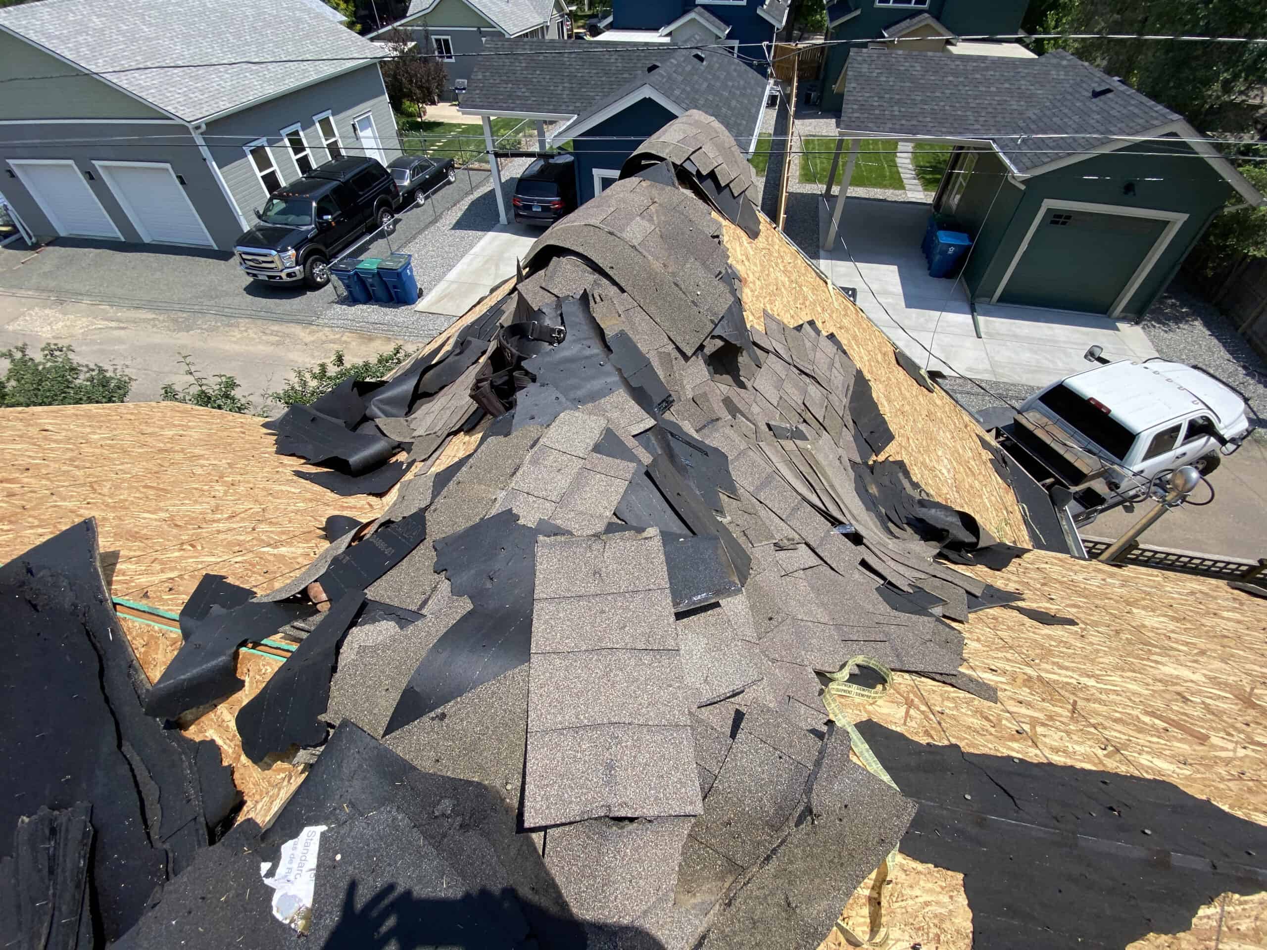 A rooftop under repair with shingles and underlayment partially removed, exposing the wooden deck. Surrounding houses and parked vehicles are visible in the background on a sunny day.