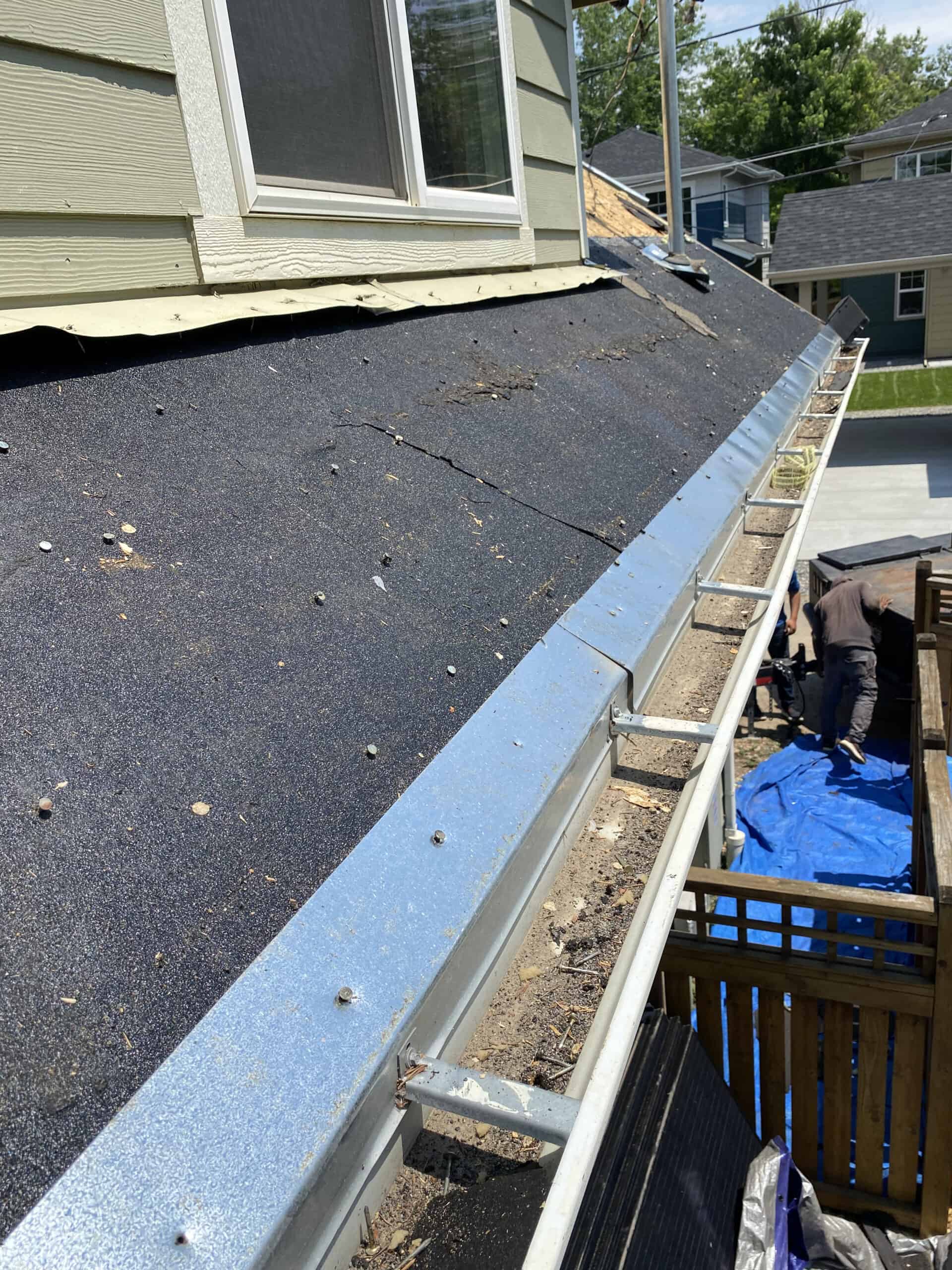 Close-up view of a house roof under repair, showing exposed roofing felt, detached gutters, and a person working below on a blue tarp. The weather appears sunny with houses and trees in the background.
