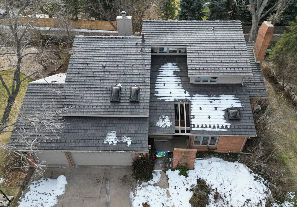 Aerial view of a two-story house with a dark shingled roof, patches of melting snow, and a brick exterior. Bare trees and light snow cover the yard and driveway, indicating a winter or early spring setting.