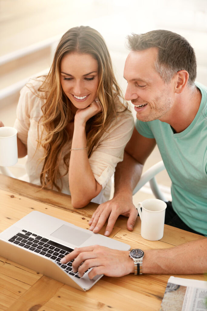 A smiling man and woman sit at a wooden table with white mugs, looking at a laptop together. The woman holds her mug, while the man points at the laptop screen. They appear relaxed and engaged.