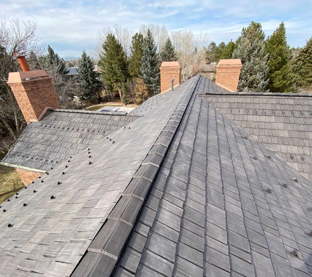 View from the top of a house roof with gray shingles, two brick chimneys, and surrounding trees in the background under a partly cloudy sky.