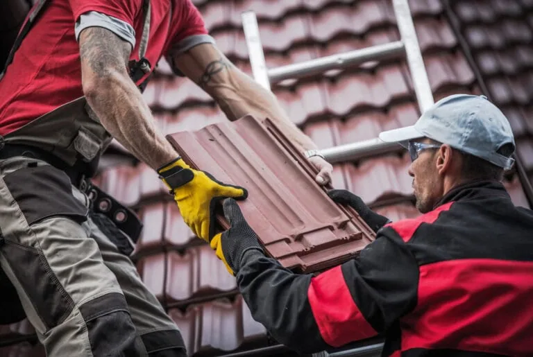 Two workers in protective clothing and gloves install brown roof tiles on a tiled roof, using a ladder for access. One man passes a tile to another who is positioned higher on the ladder, carefully fitting each piece to help ensure the longevity of the new roof—a key consideration when asking how long should a roof last.